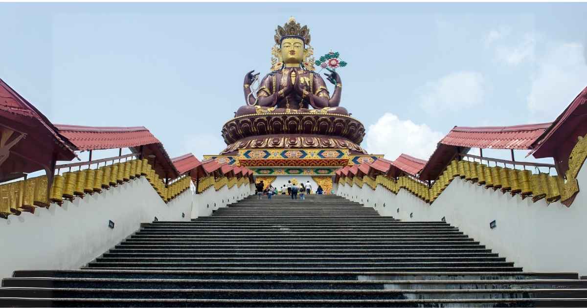 Grand staircase leading to a large golden statue of a seated deity with multiple arms, flanked by red-roofed structures.