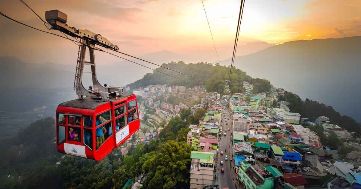 A red cable car glides over a hilly town at sunset, with colorful buildings and misty mountains in the background.