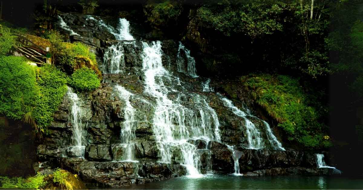 A multi-tiered waterfall cascades over dark rocky steps surrounded by dense green foliage.