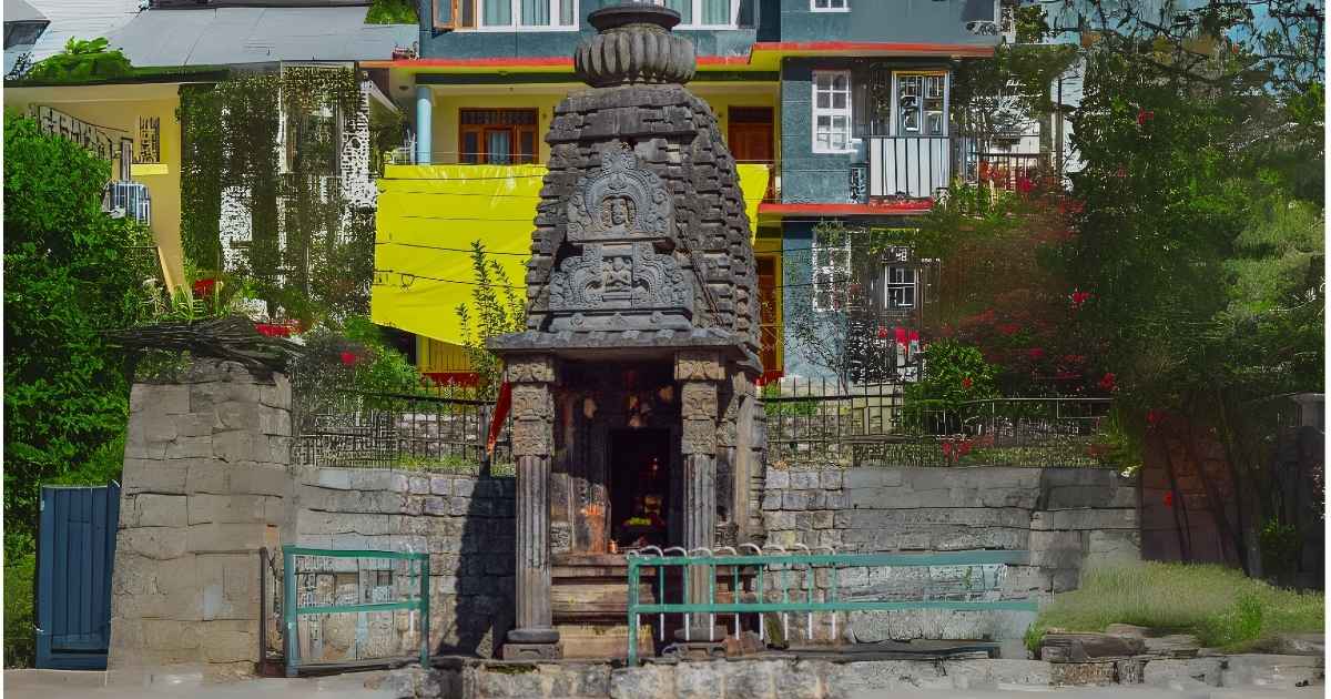 "An ancient stone temple shrine with intricate carvings, set against the backdrop of modern colorful houses in Himachal Pradesh, India."