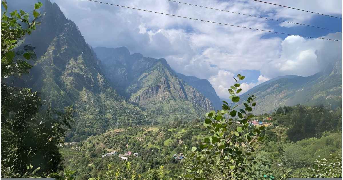 "A scenic view of a lush green valley surrounded by towering rocky mountains and scattered village houses, under a partly cloudy sky in Himachal Pradesh, India."